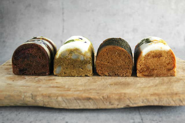 各種の色味と素材を見せる断面。黒糖の濃色、緑茶の緑色、桜島小みかんの橙色、すももを混ぜた深いココア色が鮮やかに広がるプレゼント向け蒸し菓子