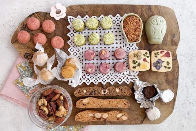 「焼き菓子ギフト（L）」のお菓子が木製ボードに美しく並べられた写真。 緑茶や紫芋味のサブレの周りには、ナッツやフクロウ型のフロランタン、エディブルフラワーとハーブがトッピングされた米粉サブレ、 キャラメル包みのアマレッティ、アルミで包まれたラム酒のベイクドショコラを配置しています。