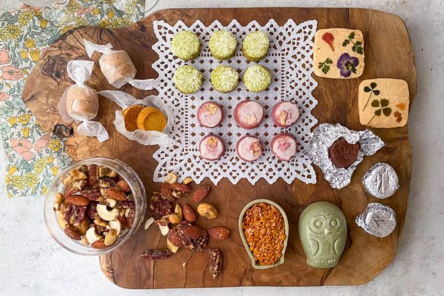 「焼き菓子ギフト（L）」のお菓子が木製ボードに美しく並べられた写真。 緑茶や紫芋味のサブレの周りには、ナッツやフクロウ型のフロランタン、エディブルフラワーとハーブがトッピングされた米粉サブレ、 キャラメル包みのアマレッティ、アルミで包まれたラム酒のベイクドショコラを配置しています。