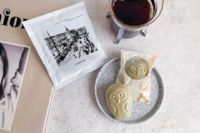 FUKU+RE(フクレ)のドリップコーヒーと、フクロウ型のフロランタンが並ぶおしゃれな朝のテーブル。コーヒーはカップに注がれており、パッケージには100年前の鹿児島の街並みが描かれている。