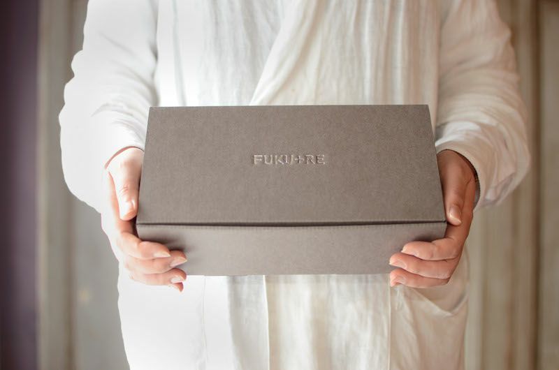 FUKU+RE(フクレ)の店員が、FUKU+REと刻印された上品なグレーのギフトボックスを両手で丁寧に持っている様子。贈り物としての丁寧さや温かみが伝わるシーン