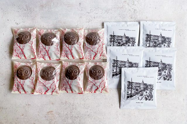 FUKU+RE(フクレ)のチョコレート味のフクロウ最中のフロランタン8個と、100年前の籠島市街地の街並みが描かれたドリップコーヒー5袋の詰め合わせ。シンプルな背景の上に整然と並べられている