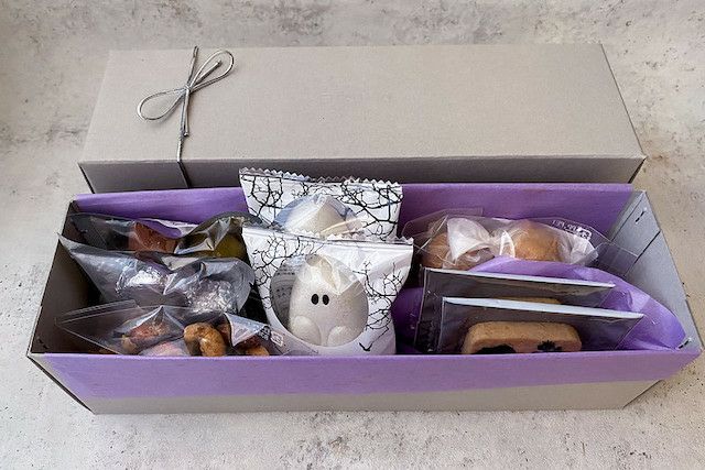 「おばけギフト（M）」のスイーツが美しくギフトボックスに詰め込まれた写真。 ラベンダーカラーの紙を敷いたギフトボックスの中には、中央にゴースト型のハロウィン限定フロランタン、両サイドにはサブレやアマレッティなどが配置されています。 ギフトボックスはグレーのシンプルなデザインです。