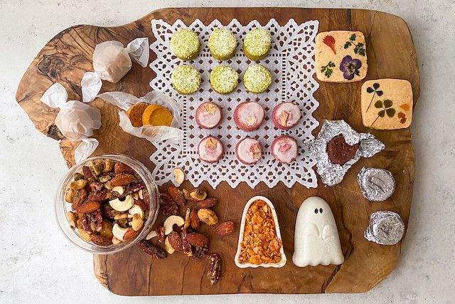 「おばけギフト（L）」のハロウィン限定スイーツが木製ボードに美しく並べられた写真。 緑茶や紫芋味のサブレの周りには、ナッツやゴースト型のフロランタン、エディブルフラワーとハーブがトッピングされた米粉サブレ、 キャラメル包みのアマレッティ、アルミで包まれたラム酒のベイクドショコラを配置しています。
